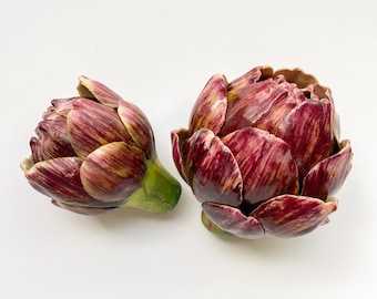 Puede incluir: Dos alcachofas, una con el tallo y las hojas intactas, la otra con el tallo quitado, ambas de color morado oscuro y verde.