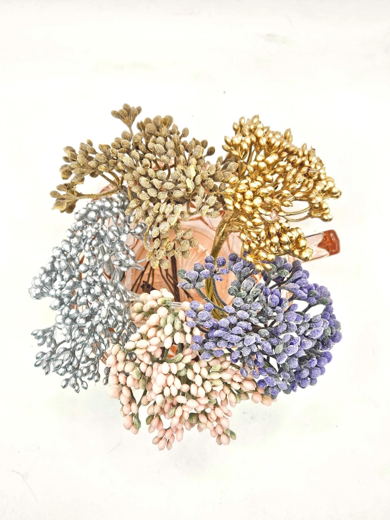 Puede incluir: Un conjunto de ramas florales artificiales en un recipiente de vidrio rosa. Las ramas presentan racimos de peque&ntilde;as bayas texturizadas en oro met&aacute;lico, plata, rosa claro, lavanda y bronce mate, dispuestas en un ramo circular.