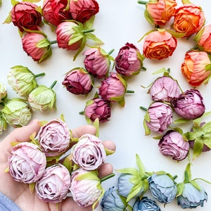 May include: Assortment of artificial flower buds in various colors, including pink, purple, blue, green, and orange. The buds are arranged on a white surface and some are held in a hand.