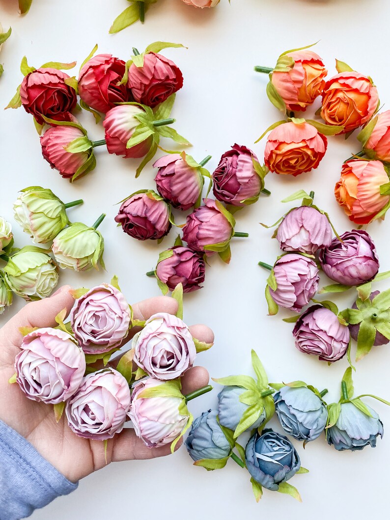 May include: Assortment of artificial flower buds in various colors, including pink, purple, blue, green, and orange. The buds are arranged on a white surface and some are held in a hand.
