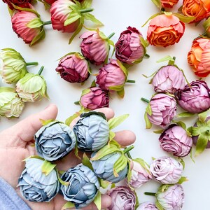 May include: Assortment of artificial flower buds in various colors, including pink, purple, orange, and green. The buds are arranged on a white surface. A hand is holding a group of blue flower buds.