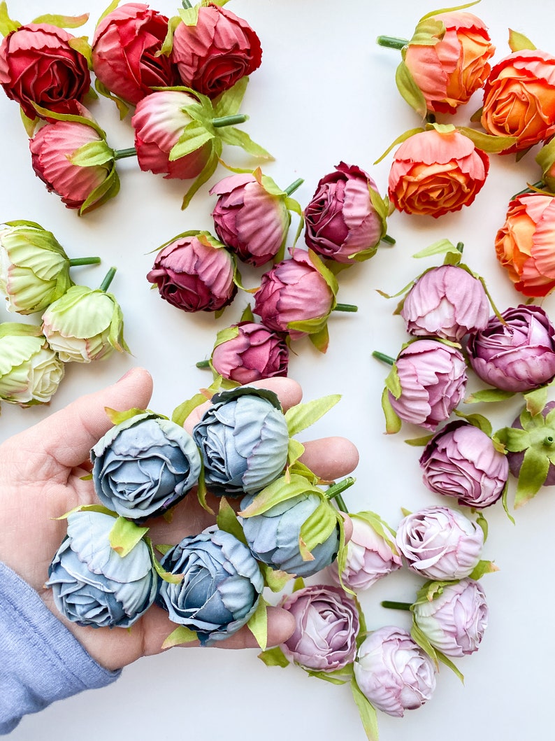 May include: Assortment of artificial flower buds in various colors, including pink, purple, orange, and green. The buds are arranged on a white surface. A hand is holding a group of blue flower buds.