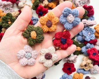 Puede incluir: Una mano sosteniendo una variedad de pequeñas flores de ganchillo en diferentes colores, incluyendo rojo, azul, verde, amarillo y marrón. Las flores están dispuestas en un círculo alrededor de la mano.