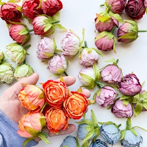 May include: Assortment of artificial flower heads in various colors, including pink, red, orange, and blue. The flowers are arranged on a white surface.