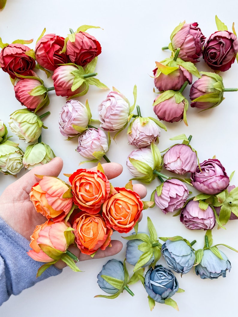 May include: Assortment of artificial flower heads in various colors, including pink, red, orange, and blue. The flowers are arranged on a white surface.