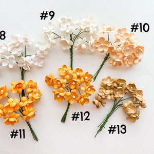 Op de afbeelding: Tien bosjes kleine papieren bloemen in verschillende kleuren, waaronder wit, perzik, geel en bruin. Elk bosje is genummerd van 8 tot en met 13.