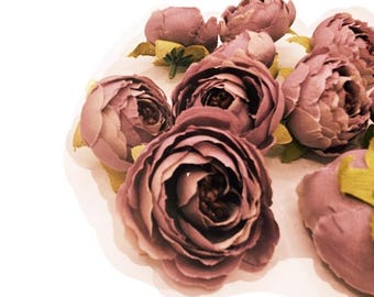 May include: A collection of mauve-colored artificial peony flowers. The blooms are in various stages of opening, with detailed petals and green and yellow accents. The flowers are arranged on a white surface, creating a soft, romantic aesthetic.