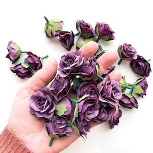 May include: A hand holding a bunch of small purple artificial roses with green stems and leaves.