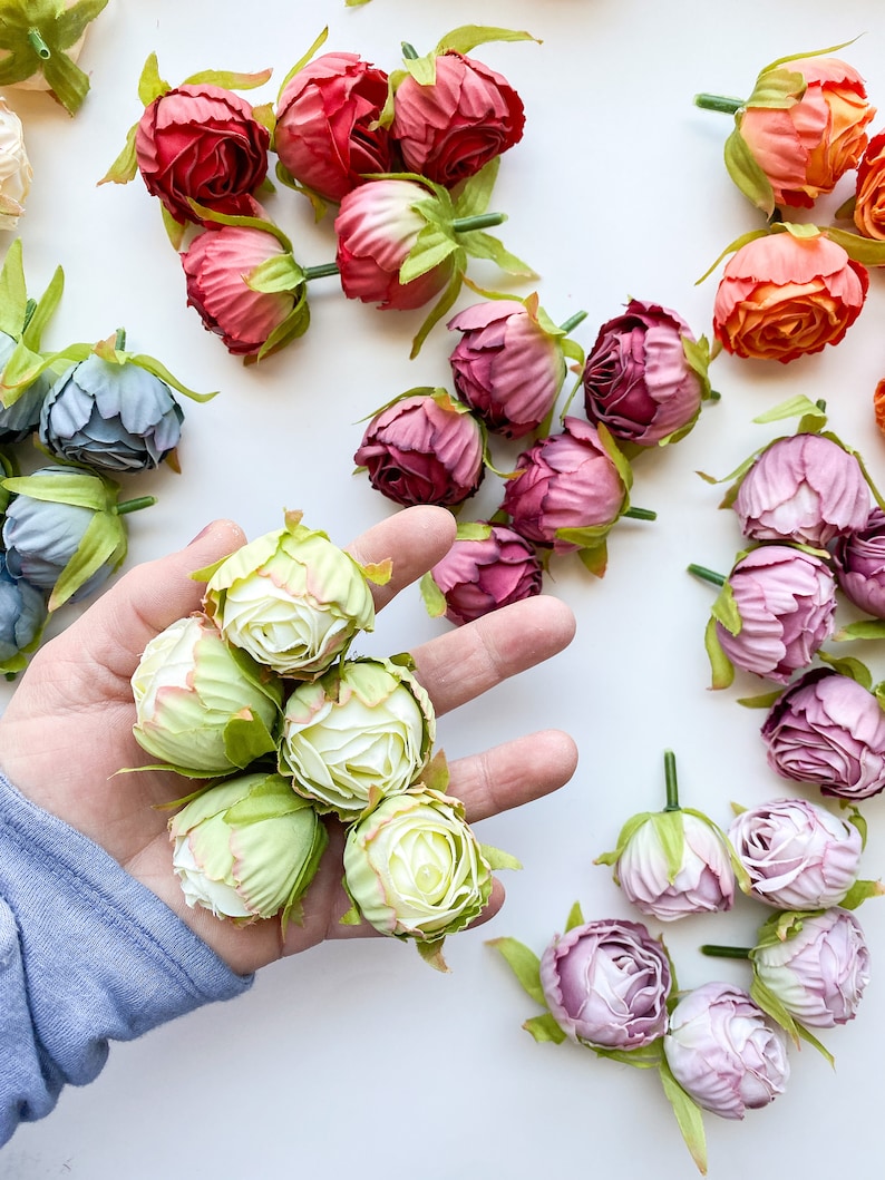 May include: A collection of artificial flower buds in various colors, including pink, red, orange, green, and blue. The buds are arranged in a hand, with the green buds in the center.