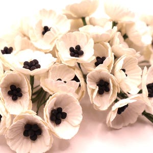 May include: A cluster of ivory-colored paper flowers with dark brown centers. The flowers have multiple petals and are arranged in a bouquet. The stems are green. These artificial flowers are suitable for crafts and decorations.