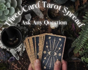 May include: A deck of tarot cards with a blue and tan design is held in two hands. The card backs feature an eye symbol with radiating lines, swords, and skulls. The text "Three Card Tarot Spread Ask Any Question" is displayed in white.