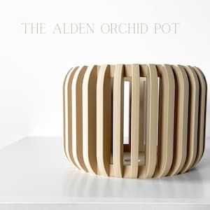 May include: A beige orchid pot made of woven wood slats. The pot is cylindrical and has a decorative design. The pot is called "The Alden Orchid Pot".