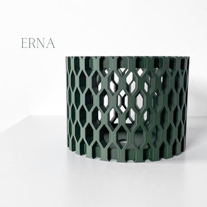 May include: A dark green cylindrical decorative object with a geometric honeycomb pattern. The word "ERNA" is printed in a light gray font on the upper left side. The object is on a white surface.