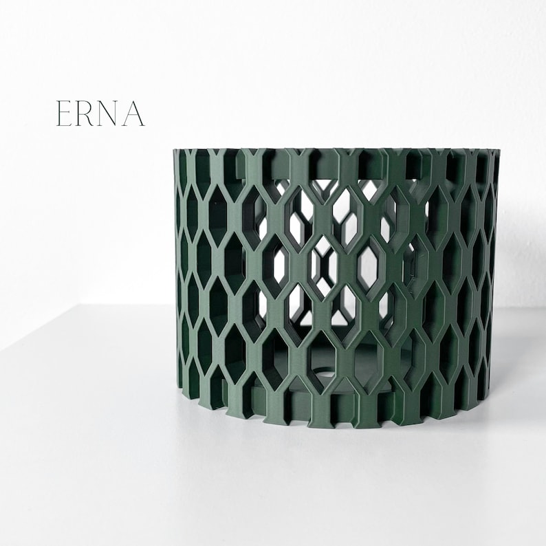 May include: A dark green cylindrical decorative object with a geometric honeycomb pattern. The word "ERNA" is printed in a light gray font on the upper left side. The object is on a white surface.