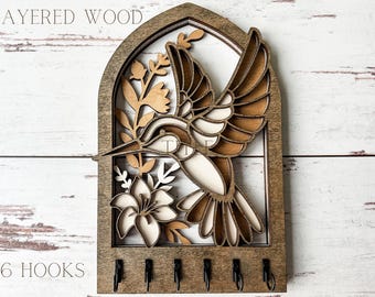 May include: Wooden key holder with a hummingbird and floral design. The key holder has six hooks and the text "LAYERED WOOD" and "6 HOOKS" is printed on the wood.