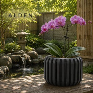 May include: A potted orchid with vibrant pink flowers and green leaves sits in a gray, vertically striped planter. The word "ALDEN" is displayed in the upper left corner. The background features a serene outdoor setting with a small waterfall and lush greenery.