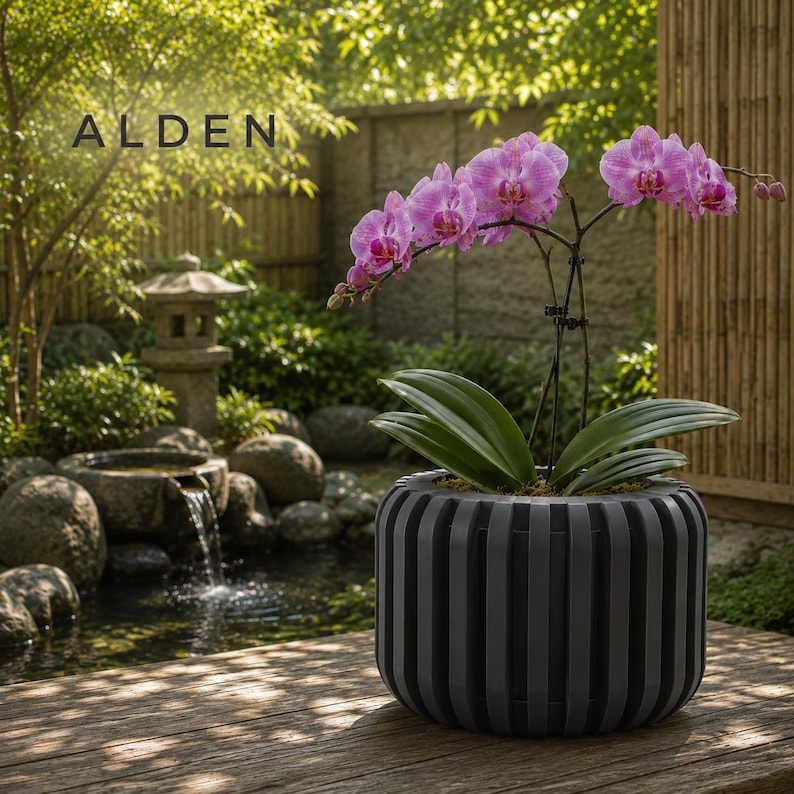 May include: A potted orchid with vibrant pink flowers and green leaves sits in a gray, vertically striped planter. The word "ALDEN" is displayed in the upper left corner. The background features a serene outdoor setting with a small waterfall and lush greenery.