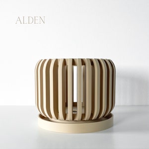 May include: A beige, cylindrical planter with vertical wooden slats, sitting on a matching saucer. The word "ALDEN" is printed in a light brown font at the top left corner. The planter has a modern, minimalist design.
