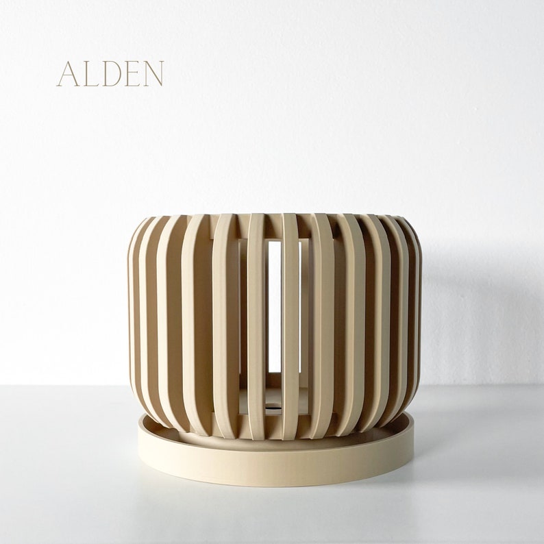 May include: A beige, cylindrical planter with vertical wooden slats, sitting on a matching saucer. The word "ALDEN" is printed in a light brown font at the top left corner. The planter has a modern, minimalist design.