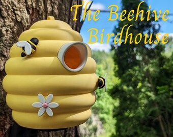 May include: A yellow beehive-shaped birdhouse with the text "The Beehive Birdhouse". The birdhouse has a round entrance and is decorated with a white and pink flower and two cartoon bees. The birdhouse is mounted on a tree trunk.