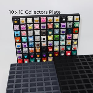 May include: A black 10 x 10 collectors plate with a grid of 100 colorful mechanical keyboard keycaps. The plate is made of plastic and has a black background. The keycaps are arranged in rows and columns, and each keycap has a different color and design.