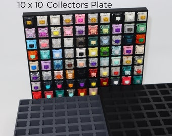 May include: A black 10 x 10 collectors plate with a grid of 100 colorful mechanical keyboard keycaps. The plate is made of plastic and has a black background. The keycaps are arranged in rows and columns, and each keycap has a different color and design.