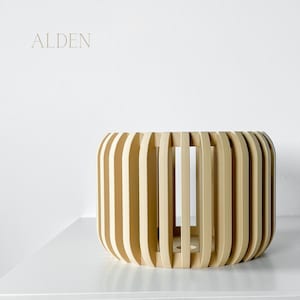 May include: A light beige, cylindrical wooden planter with vertical slats. The planter has a rounded shape and sits on a white surface. The word "ALDEN" is printed in a light brown color in the upper left corner.