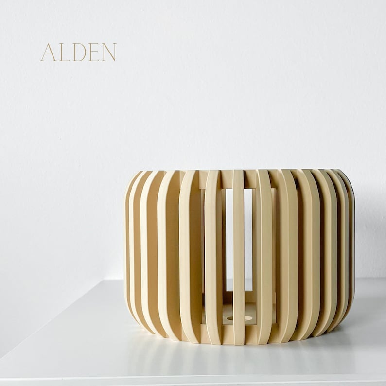 May include: A light beige, cylindrical wooden planter with vertical slats. The planter has a rounded shape and sits on a white surface. The word "ALDEN" is printed in a light brown color in the upper left corner.