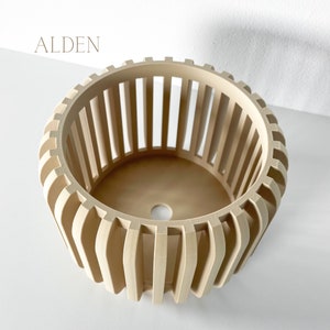 May include: A beige, cylindrical planter with vertical slats and a circular base. The word "ALDEN" is printed in a light brown font at the top left. The planter has a modern, geometric design.