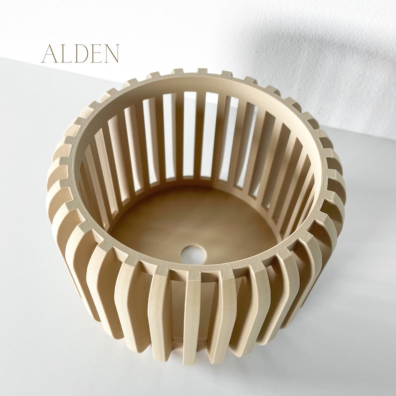 May include: A beige, cylindrical planter with vertical slats and a circular base. The word "ALDEN" is printed in a light brown font at the top left. The planter has a modern, geometric design.