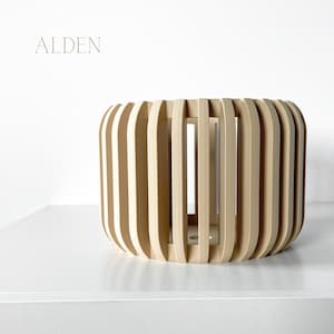 May include: A cylindrical wooden decorative object with vertical slats. The object is light-colored and has the word "ALDEN" in a light brown font at the top left. The object is sitting on a white surface.