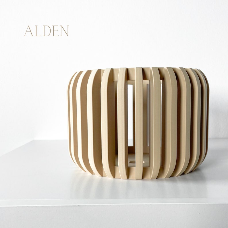 May include: A cylindrical wooden decorative object with vertical slats. The object is light-colored and has the word "ALDEN" in a light brown font at the top left. The object is sitting on a white surface.