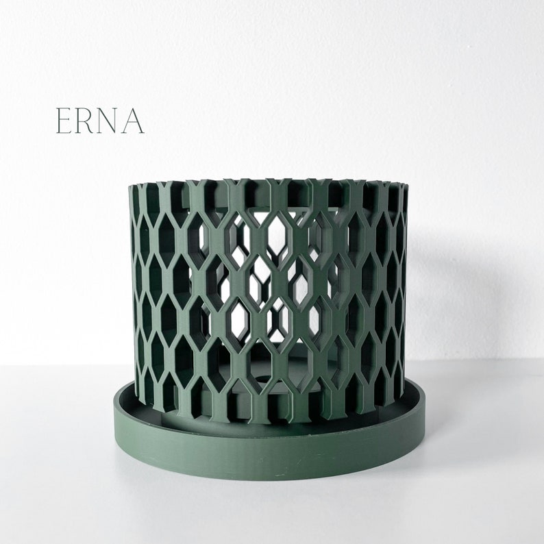 May include: A dark green cylindrical planter with a geometric honeycomb design. The planter sits on a matching round base. The word "ERNA" is printed in a light gray font on the upper left side of the image.