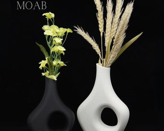 May include: Two modern vases, one black and one white, each with a circular opening. The black vase holds yellow flowers, while the white vase contains wheat stalks. The word "MOAB" is printed in white on the upper left.