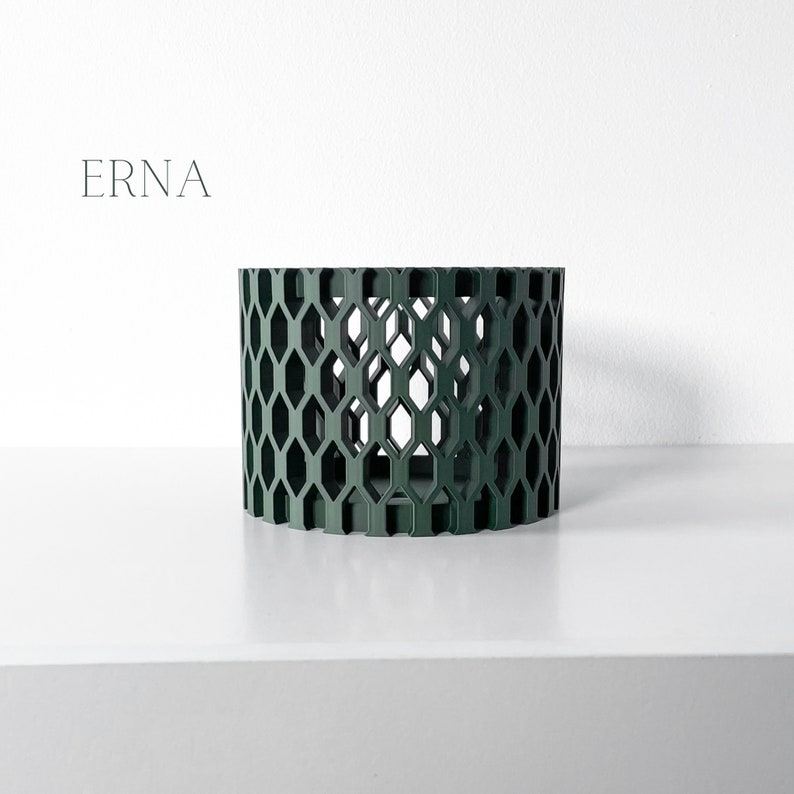 May include: A dark green cylindrical decorative object with a geometric honeycomb pattern. The object is on a white surface against a white background. The word "ERNA" is printed in a minimalist font on the upper left.