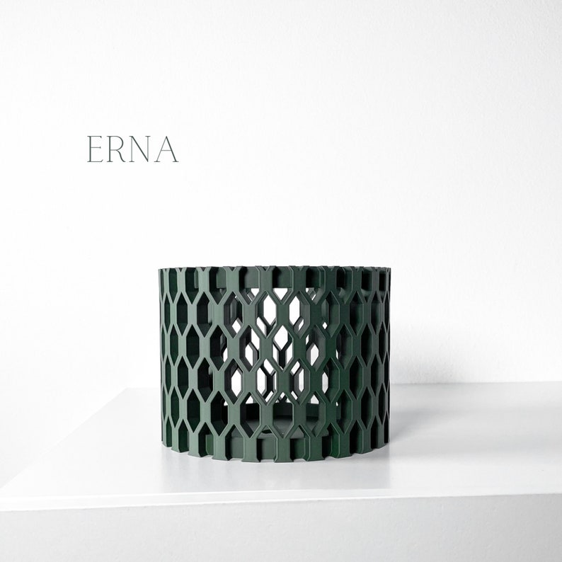 May include: A cylindrical, dark green decorative object with a geometric honeycomb pattern. The word "ERNA" is printed in a minimalist font on the white background. The object is placed on a white surface.