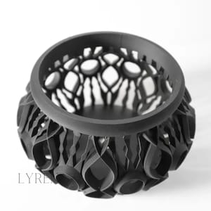 May include: A black, decorative bowl with a unique, openwork design. The bowl features a circular rim and intricate cut-out patterns, creating a visually appealing texture. The base has a series of curved shapes. The word "LYREN" is printed on the side.