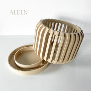 May include: A light-colored wooden planter with a matching saucer. The planter has a cylindrical shape with vertical slats, and the saucer is round. The word "ALDEN" is printed in the upper left corner.