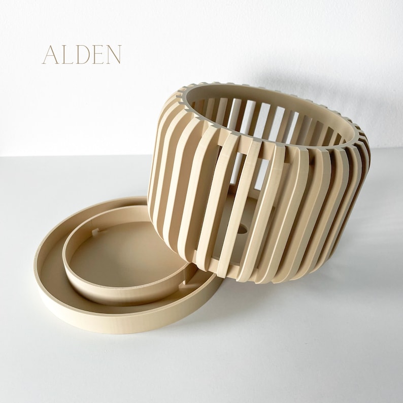 May include: A light-colored wooden planter with a matching saucer. The planter has a cylindrical shape with vertical slats, and the saucer is round. The word "ALDEN" is printed in the upper left corner.