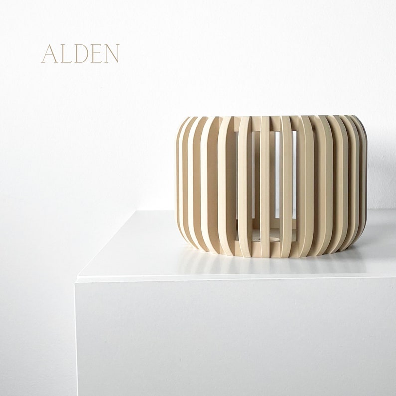 May include: A light-colored wooden decorative bowl with vertical slats, set against a white background. The word "ALDEN" is printed in a light brown color on the upper left side. The bowl is cylindrical with rounded edges.