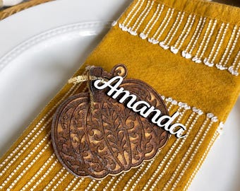 May include: A mustard yellow cloth napkin with white vertical stitched stripes, featuring a dark brown laser-cut wooden pumpkin ornament. The pumpkin has an intricate filigree pattern and a white script name tag attached that reads Amanda.