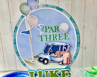 May include: A golf-themed cake topper in blue and green. The design features a golf cart, golf ball, flag, and balloons. The text "PAR THREE" and the name "LUKE" are also included.