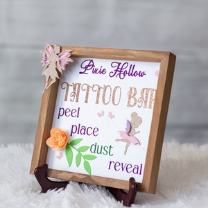 May include: A wooden sign with a glittery background and the words "Pixie Hollow Tattoo Bar" in purple lettering. The sign also includes the words "peel, place, dust, reveal" in purple lettering. The sign is decorated with a pink and purple fairy, a flower, and green leaves.