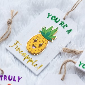 May include: A white wooden tag with a gold glitter pineapple design and the text "You're a Pineapple!" in gold glitter. The tag is tied with twine.
