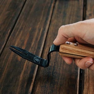 May include: A black metal weeding tool with a wooden handle. The handle has a brown leather strap and is held in a hand.