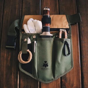 May include: A green canvas tool belt with multiple pockets and a black strap. The belt has a knife, gloves, a small saw, and other tools. The belt has a black patch with the text "OLD SOUL" and a clover logo.