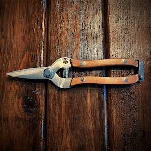 May include: A pair of gardening shears with wooden handles. The handles are brown and have a metal clasp. The blades are silver and have a small clover-shaped logo on the side. The logo says "GARDENA".