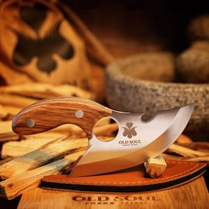 May include: A wooden handled knife with a stainless steel blade. The blade has a unique shape with a hole in the center. The knife is resting on a wooden surface with a brown leather pad. The knife has the words "Old Soul" and "Young Spirit" engraved on the blade.