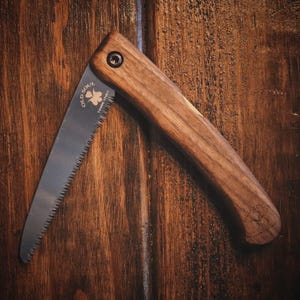 May include: A folding saw with a wooden handle. The saw blade is made of metal and has a black finish. The handle is made of dark wood and has the words "Old Soul" and "Young Spirit" engraved on it.