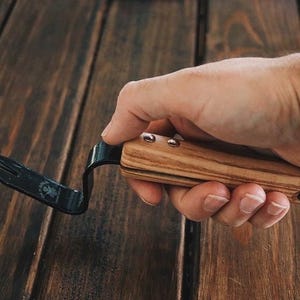 May include: A black metal hand weeding tool with a wooden handle. The handle is made of light and dark wood with a leather strap.